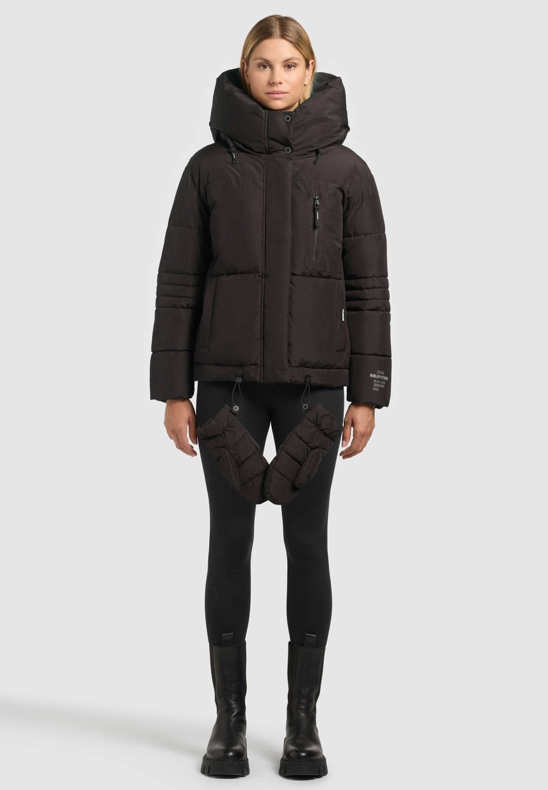 Doudoune noire matelassée avec une grande capuche, une poche poitrine zippée et un ourlet à cordon de serrage. Comprend des accents matelassés assortis sur les jambes. Portée avec des leggings noirs et des bottes.