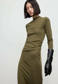 Abito aderente verde oliva con maniche lunghe, dettaglio arricciato laterale e maglia testurizzata. Abbinato a un guanto di pelle nera che si estende fino all'avambraccio.