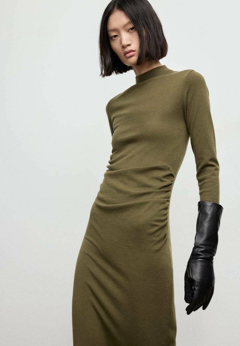 Abito aderente verde oliva con maniche lunghe, dettaglio arricciato laterale e maglia testurizzata. Abbinato a un guanto di pelle nera che si estende fino all'avambraccio.