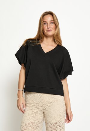 Giovane donna con lunghi capelli biondi che indossa un top nero con scollo a V e pantaloni beige in pizzo, in piedi davanti a uno sfondo bianco semplice.