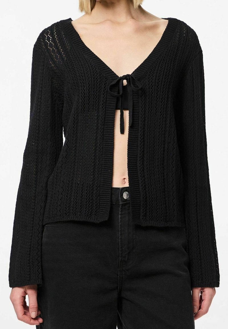 Femme portant un cardigan en tricot noir noué sur la poitrine, avec des manches longues et un pantalon noir taille haute.