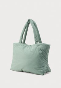 Απαλή, επενδυμένη, ανοιχτοπράσινη τσάντα tote με δύο χοντρές λαβές, ορθογώνιο σχήμα και καπιτονέ ραφές σε απλό ανοιχτό φόντο.