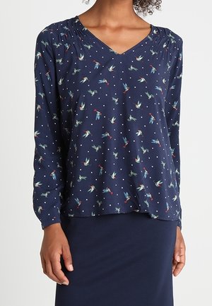Vrouw draagt een marineblauwe blouse met lange mouwen met kleine vogel- en polkadotprints en een bijpassende marineblauwe rok.