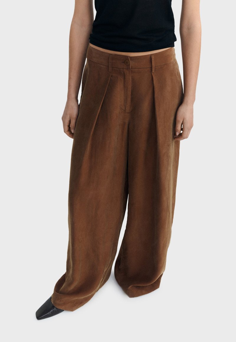 Bruine wijdvallende broek met plooien gedragen door persoon in zwarte mouwloze top en zwarte puntige schoenen op witte achtergrond.