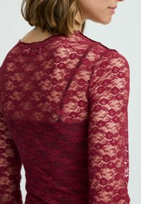 Top in pizzo borgogna con maniche lunghe trasparenti e motivo floreale. La schiena è parzialmente foderata, mettendo in mostra dettagli in pizzo intricati.
