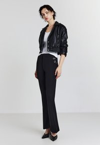 Schwarze cropped Lederjacke mit Druckknöpfen, weißes Tanktop, figurbetonte schwarze Hose mit silbernen Ringakzenten und schwarze spitze High Heels.