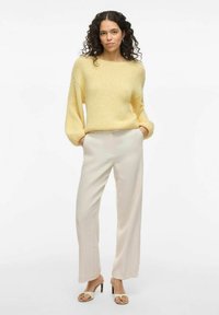 Rumena pletena jopica s širokimi rokavi, v kompletu s kremnimi širokimi hlačami. Model nosi sandale s peto. Preprost ozadje.