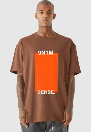 Barna pamut póló feltűnő narancssárga téglalap alakú grafikával, amelyen fehér betűkkel a "9N1M SENSE" felirat szerepel. Rövid ujjú, laza szabású.