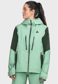 Frau mit dunklem Pferdeschwanz, die eine mintgrüne und dunkelgrüne Kapuzenjacke für den Außenbereich mit Reißverschlusstaschen trägt und vor einem hellen Hintergrund steht.