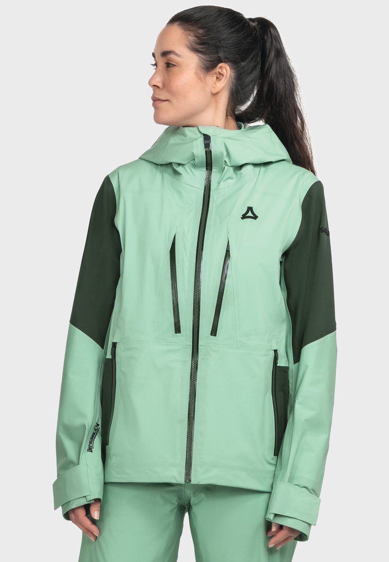 Frau mit dunklem Pferdeschwanz, die eine mintgrüne und dunkelgrüne Kapuzenjacke für den Außenbereich mit Reißverschlusstaschen trägt und vor einem hellen Hintergrund steht.