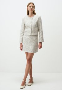 Kurze Tweedjacke mit rundem Ausschnitt und vorderen Knöpfen, kombiniert mit einem passenden Minirock. Beide in Creme mit blauen Punkten. Schuhe mit quadratischem Absatz.