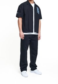 Camisa negra de manga corta con cuello y letras azules bordadas, combinada con pantalones negros y zapatillas blancas. Tela suave, corte relajado.