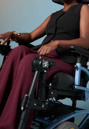 Person iført en ærmeløs sort top og vinrøde bukser, siddende i en blå elektrisk kørestol mod en ensfarvet baggrund.