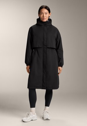 Black waterproof long coat with a cropped design, high collar, elastic cuffs, and side pockets, worn over black leggings and white sneakers.