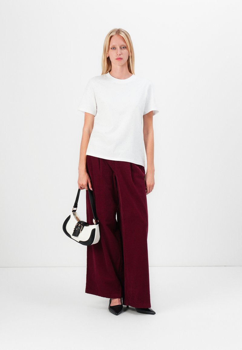 T-shirt bianca a maniche corte, abbinata a pantaloni larghi bordeaux. Il modello tiene una borsa in bianco e nero e indossa scarpe nere a punta.