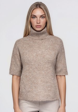 Une femme porte un pull en maille à col roulé et à manches courtes, de couleur taupe, sur un fond uni et clair.