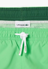 Groene zwemshorts met een elastische tailleband, witte trekkoord en mesh voering. Bevat een Lacoste-logo label op de taille.