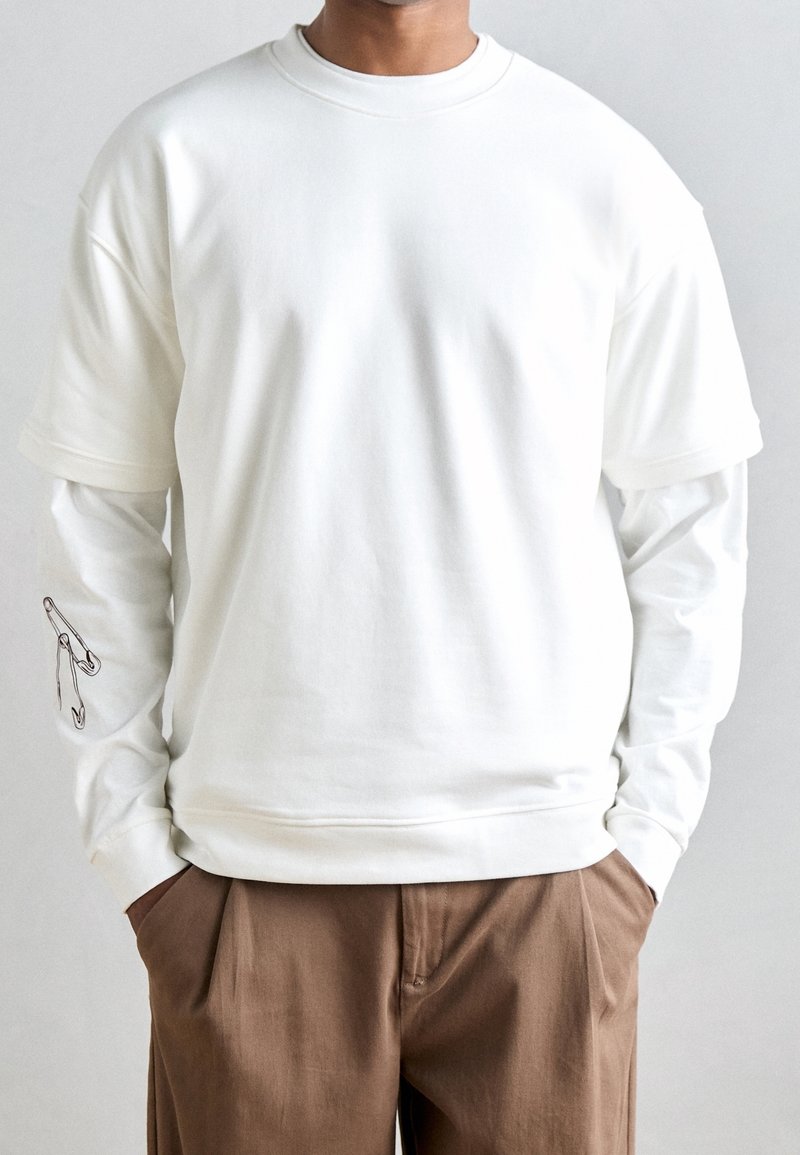 Personne portant des chemises blanches à manches longues et courtes superposées avec des motifs d'épingles de sûreté sur la manche, associées à un pantalon marron, mains dans les poches.