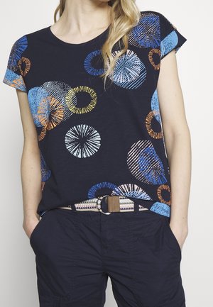 Nainen pukeutuneena tummansiniseen paitaan, jossa värikkäitä pyöreitä kuvioita, ja tummansinisiin housuihin, joissa on beige kudottu vyö, kädet taskuissa.