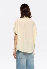 Short-sleeve, loose-fitting shirt in light yellow textured fabric, featuring a curved hem and a simple collar. Back view shows relaxed fit.
