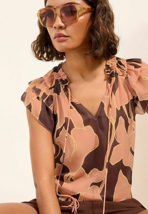 Lühikeste laineliste juustega naine, kes kannab ülikõrgeid merevaiguvärvi päikeseprille ning pruuni ja virsikuvärvilise lillelise varrukateta pluusi voldikute jaotusega õlgadel.