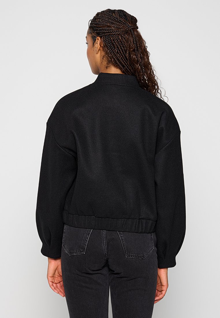 Schwarze Bomberjacke mit hohem Kragen, fallenden Schultern und elastischem Saum. Glattes Material und entspannter Schnitt mit gefalteten Ärmeldetails.