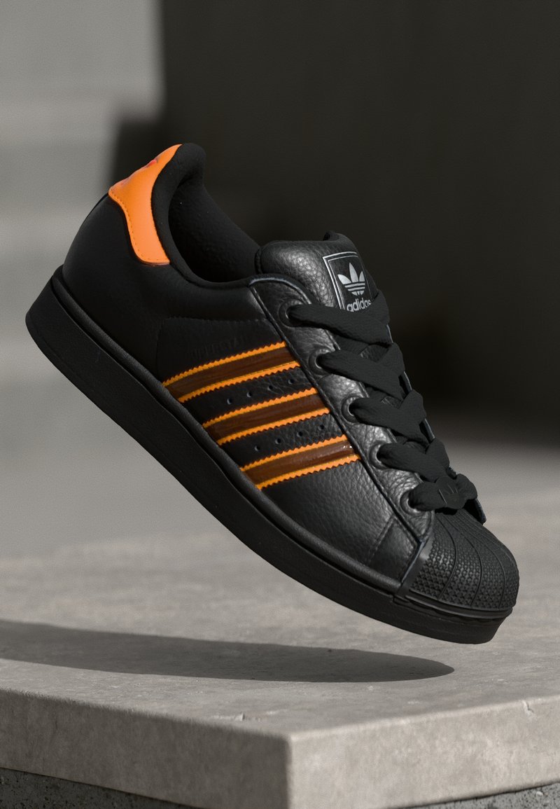 Adidași negri cu accente portocalii, trei dungi orizontale, parte superioară din piele texturată, capac de cauciuc pentru vârf și șireturi negre.