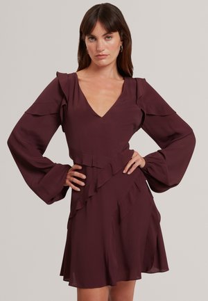 Femme aux longs cheveux foncés porte une robe violet foncé avec de longues manches bouffantes, des volants aux épaules et à la jupe, debout les mains sur les hanches.