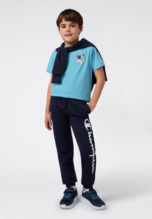 Ragazzo che indossa una maglietta blu con grafica di un astronauta, pantaloni blu scuro con grande testo bianco "Champion", sneakers abbinate e una felpa blu scuro appoggiata sulle spalle.