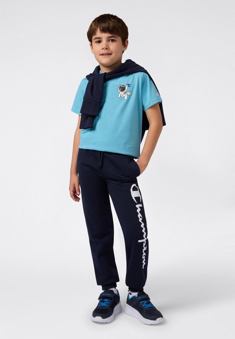 Niño con una camiseta azul con un gráfico de astronauta, pantalones marinos con un gran texto blanco "Champion", zapatillas a juego y una sudadera marina caída sobre los hombros.