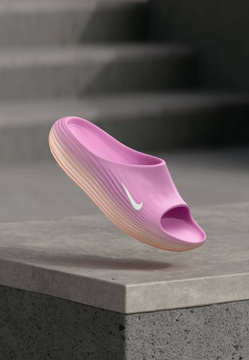 Roza Nike papuča s bijelim swoosh logotipom, teksturiranim potplatom i otvorenim prstima lebdi iznad betonske površine.