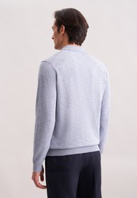 Mann mit dunklem Haar trägt einen hellgrauen Langarmpullover und dunkle Hosen, steht mit dem Rücken zur Kamera vor einem schlichten Hintergrund.