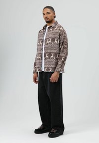 Veste chemise marron à motifs avec des cerfs et des arbres, fermeture à boutons, associée à un pantalon noir à jambes larges et des chaussures noires avec des lacets roses.