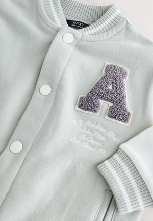 Vaaleanharmaa urheilutakki valkoisilla napeilla, raidoitettu ribbineuloskaulus, violetti fleece-kirjain "A" -merkki ja kirjailtuja yksityiskohtia rinnassa.