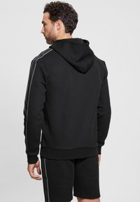 Black hooded sweatshirt with white side stripes, made of soft fabric. Features a kangaroo pocket and ribbed cuffs, paired with shorts.