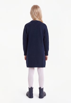 Kind met lang blond haar, gekleed in een marineblauwe jurk met lange mouwen, witte panty's en donkere laarzen, staand met de rug naar de kijker op een witte achtergrond.