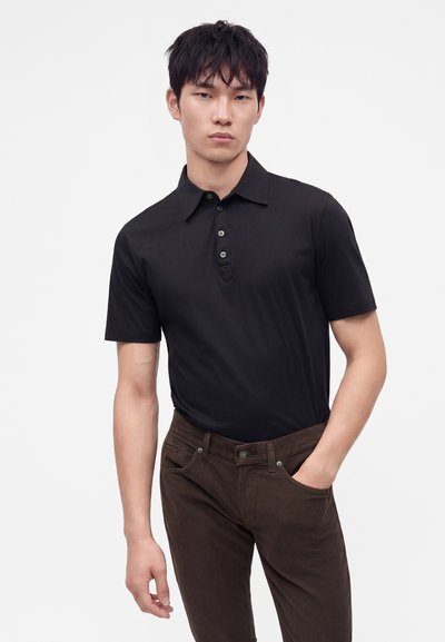Jeune homme debout portant un polo noir à manches courtes et un pantalon marron foncé, une main dans la poche.