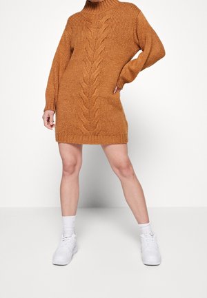Femme portant une robe-pull en maille torsadée marron à manches longues, associée à des chaussettes blanches et des baskets blanches, debout les jambes écartées.