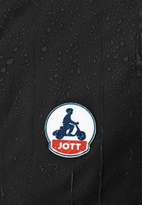 Toppa circolare JOTT con design di uno scooter, blu su sfondo bianco con base rossa, posizionata su una superficie nera testurizzata con gocce.