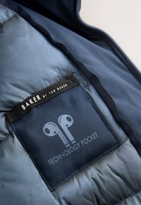 Indersiden af en marineblå polstret jakke, der viser en etiket med teksten "Technology Pocket" med et ikon af en øretelefon. Stoffet er glat med en let glans.