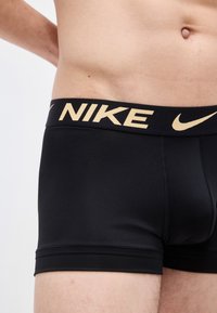 Torso masculin portant un boxer Nike noir avec logo beige sur la ceinture, montrant la peau de l'ombilic jusqu'au milieu de la cuisse sur un fond uni.