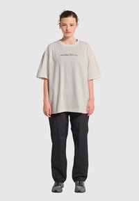 Przesadny beżowy bawełniany t-shirt z krótkimi rękawami, z czarnym napisem i logo. Połączony z czarnymi spodniami i szarymi trampkami.