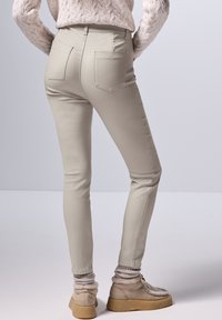 Beige skinny pants met achterzakken, gecombineerd met grijze gebreide sokken en beige laarzen, tegen een lichtgrijze achtergrond.