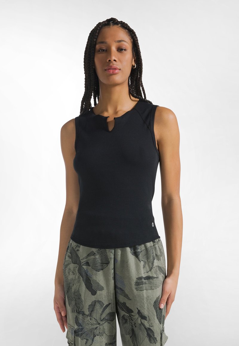 Mujer joven con el cabello trenzado, vestida con una camiseta sin mangas negra y pantalones cargo verde con estampado de camuflaje, sobre un fondo blanco liso.