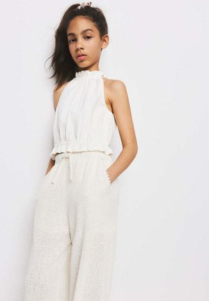 Fille aux cheveux foncés attachés en arrière, portant un haut blanc sans manches à volants et un pantalon crème texturé taille haute, les mains dans les poches.