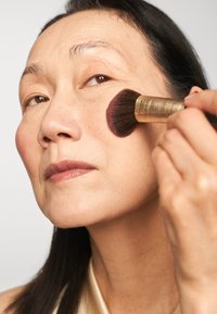 Pennello per illuminante con setole grandi e morbide che applica il fard su un incarnato chiaro. Il soggetto ha capelli scuri e lisci e un finish naturale.