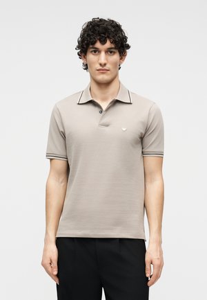 Jeune homme aux cheveux bouclés portant un polo beige à manches courtes et un pantalon noir, debout devant un fond clair uni.