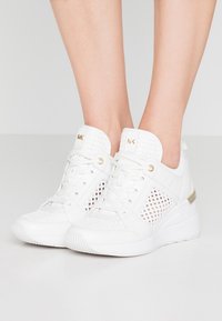 Sneakers sportivi bianchi con dettagli in rete, accenti dorati e tacco a zeppa. Scamosciato sintetico testurizzato e design della suola a contrasto.