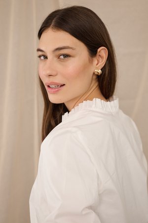 Fiatal nő hosszú barna hajjal, arany karika fülbevalóval és fodros galléros fehér blúzzal, aki hátrafordulva néz.