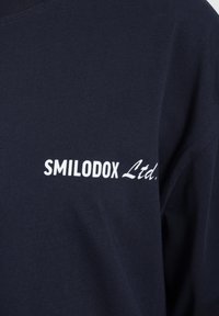 Námořnická modrá tričko s kulatým výstřihem a krátkými rukávy, na přední straně s bílým potiskem "SMILODOX Ltd." Hladký povrch.